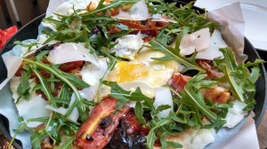 Pizza with bacon, cheese shavings, arugula or rocket, and egg in the middle